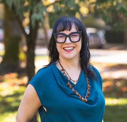 Trish Kelly, a light skinned woman with dark hair and thick black glasses.